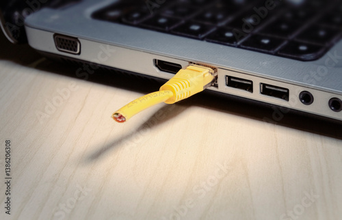 Cut yellow ethernet cable connected to laptop port, revealing exposed wires at cable end. Close-up view. Symbolic shot: sabotage, malfunction, censorship. With ominous shadows.
