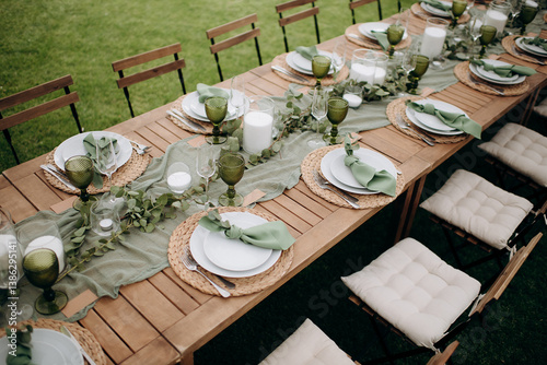 Elegant outdoor wedding table setting with a wooden table, sage green runner, white plates, and green wine glasses