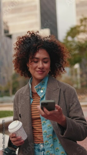 Vertical video,A young woman smiles as she walks down the street while talking on her cell phone. She is happy and confident.