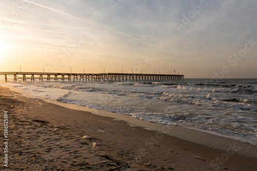 Waves crash along the shore as Oak Island Pier extends into the Atlantic Ocean under a glowing sunrise sky in North Carolina.