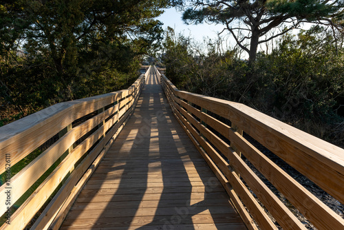 A long wooden footbridge stretches through coastal trees and marshland in Oak Island, North Carolina, under a clear blue sky.