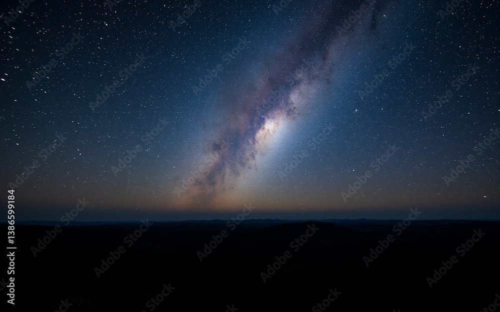 Fototapeta premium A breathtaking view of the night sky showcases the bright milky way stretching across the dark expanse above a shadowy landscape. Countless stars twinkle against the deep blue gradient.