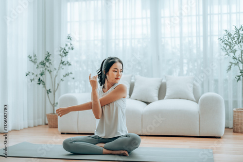 Happy young Asian woman practicing yoga and shoulder stretching at home sitting on floor in living room