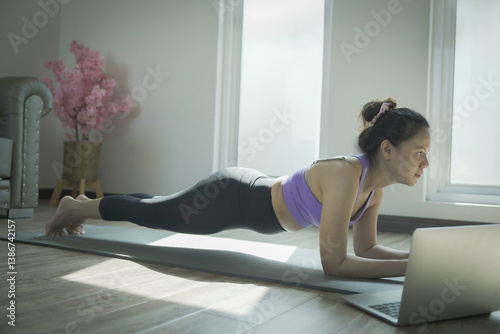 A woman is doing a plank on a mat while looking at a laptop. The laptop is open and the woman is focused on the screen