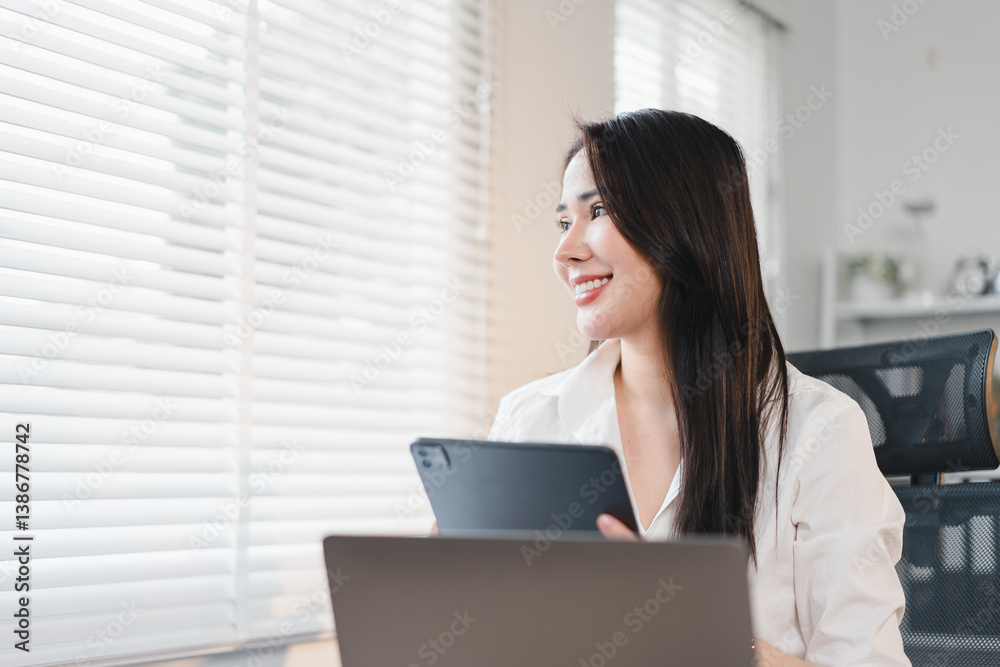 Cheerful woman in white shirt holding tablet, sitting at desk with laptop, looking out window with blinds. Cheerful woman in white shirt holding tablet, sitting at desk with laptop, looking out window with blinds.