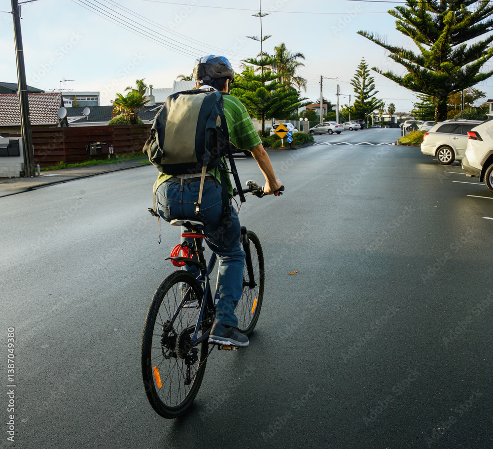 Cyclist riding on the suburb road in the morning. Houses and parked cars on the sides of the road. Auckland.