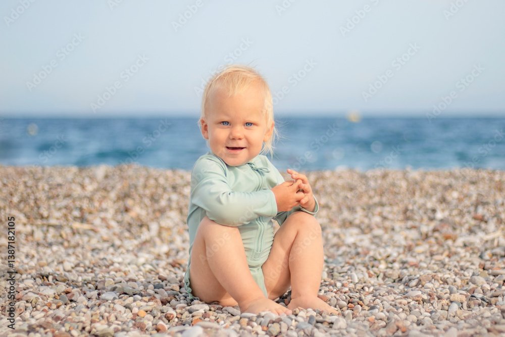 A joyful and cheerful toddler joyfully sits on a pebbly beach, smiling brightly with the vast ocean in the background