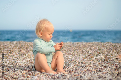Obraz na płótnie A joyful and cheerful toddler joyfully sits on a pebbly beach, smiling brightly with the vast ocean in the background w salonie