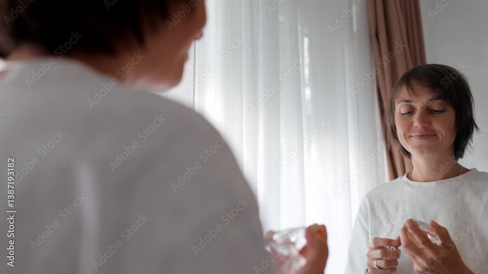 Young woman stands in front of a mirror, sniffs eau de toilette, sprays it on herself, and smiles, enjoying the aroma. Perfect for themes of beauty, fragrance, and self-care.