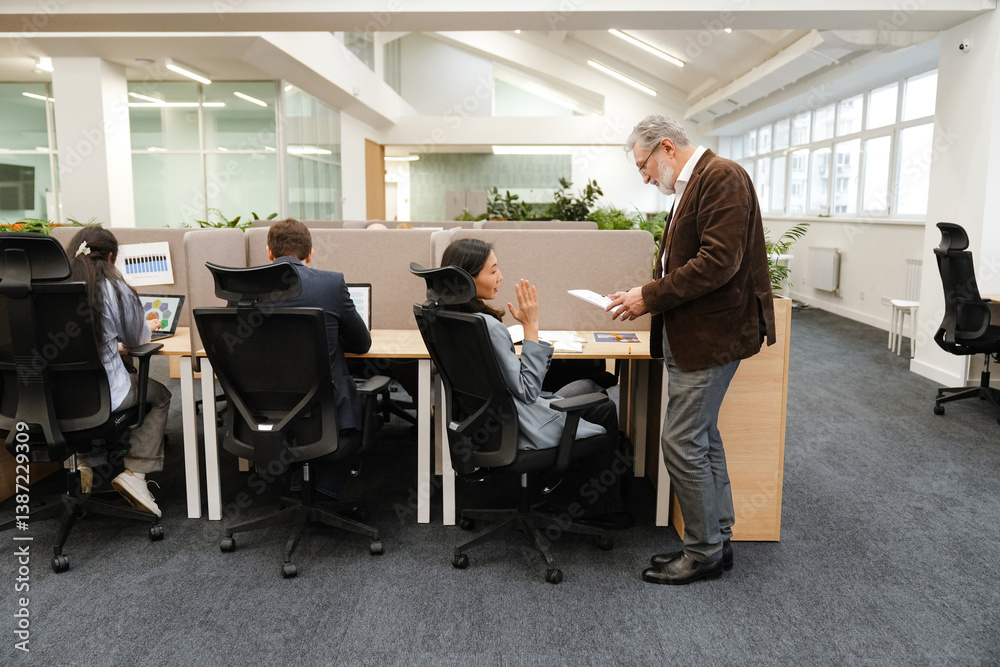 In an office, a gray-haired White man in his 60s stands and shows documents to his Asian female colleague in her 30s sitting at her desk and talking, with other workers at their desks near them. In an office, a gray-haired White man in his 60s stands and shows documents to his Asian female colleague in her 30s sitting at her desk and talking, with other workers at their desks near them.
