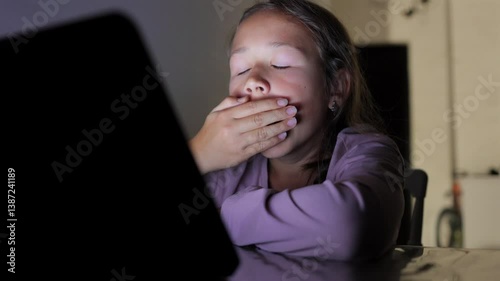 A young girl sits at a table, looking at a tablet screen. She appears tired and sleepy, yawning as she struggles to stay awake. Perfect for themes of screen time and fatigue.