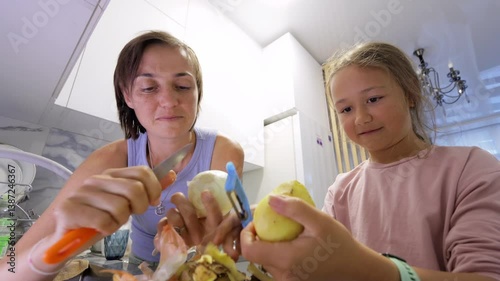 Close-up view from below of a mother and daughter in the kitchen, peeling vegetables, talking, and laughing. A warm family moment perfect for cooking or lifestyle themes.