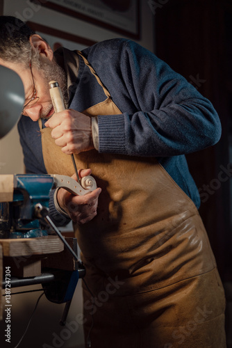 Skilled craftsman creating violin scroll, carefully carving intricate wood details using specialized gouge and chisel, securing delicate instrument part in workshop vise