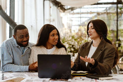 Wallpaper Mural A Black couple in their 30s sitting at a table with a laptop during a meeting with a female adult European wedding organizer in a blazer in a room with glass walls, plants, and high ceiling Torontodigital.ca