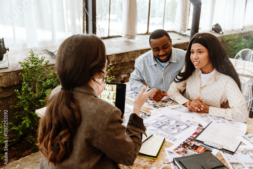 Two diverse women, and an adult Black man with short dark hair, sit at a table discussing photos and documents. Large windows and greenery are in the background. The people discuss a wedding.