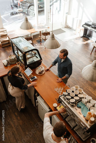 An overhead view of a mid-30s White man ordering coffee while standing in a small coffee shop, as a young, White woman in her 20s stands behind the counter, with her male colleague making the order