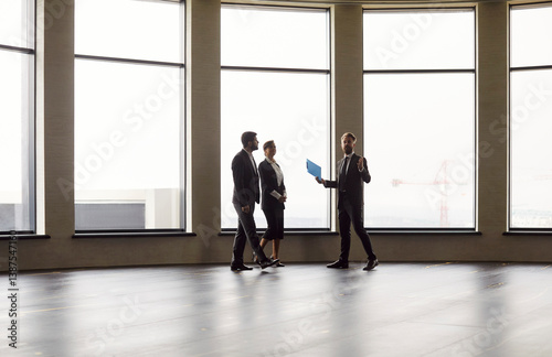 Business professionals in suits walking through spacious modern office with panoramic windows. Confident realtor presenting real estate, explaining project details to potential buyers or investors.