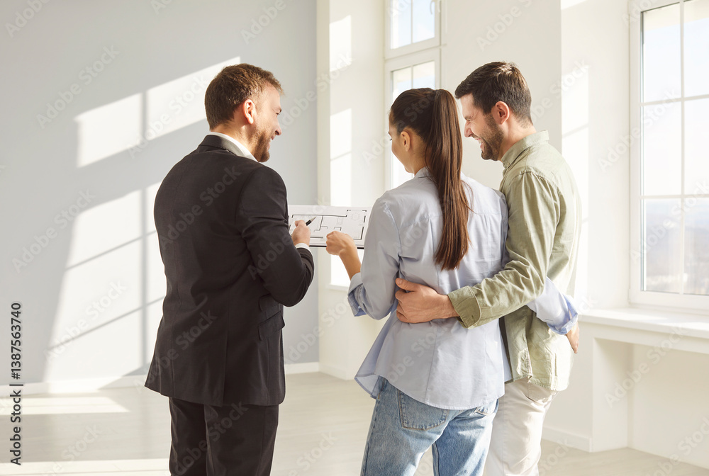 Fototapeta premium Back view of real estate agent in suit presenting apartment layout to happy young couple in bright empty room. Smiling man and woman hugging each other, discussing future home purchase with realtor.