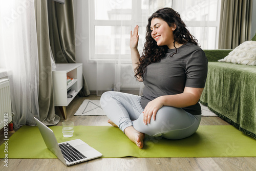 Free Download Side view of overweight young beautiful woman sitting on mat with glass of water in living-room in front of laptop, waving hand greeting her online fitness instructor, ready to start training at home - GFXLoad