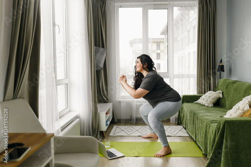 Losing weight online fitness program concept. Plus size woman in sports clothes training at home, doing squats on green mat in her living-room following instructions of her interlocutor using laptop
