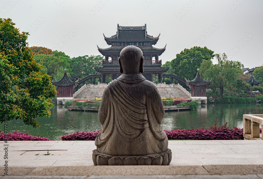 Obraz premium Serene view of a stone Buddha facing a traditional pavilion near a tranquil lake in a Chinese garden