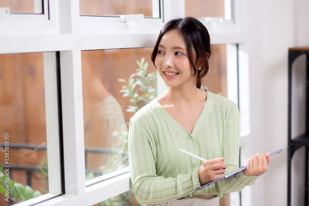 Young asian woman using tablet writing with inspiration and comfortable by window in living room at home office, cheerful woman standing working on digital tablet in workspace, business and lifestyle. Young asian woman using tablet writing with inspiration and comfortable by window in living room at home office, cheerful woman standing working on digital tablet in workspace, business and lifestyle.