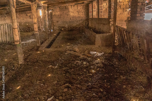 An atmospheric interior view of an old, abandoned barn or stable with rustic wooden beams, worn brick walls, and feeding troughs. The floor is covered with dirt and hay remnants
