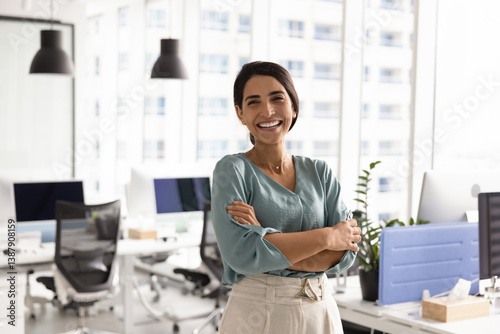 Happy successful female Latin businesswoman posing in office workspace alone, looking at camera, smiling, standing with arms crossed. Confident cheerful business leader woman portrait
