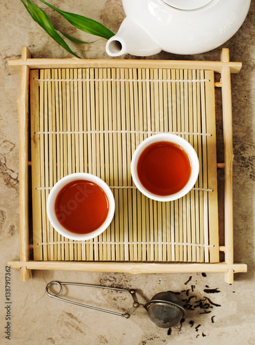 Overhead view of two cups of Chinese Pu-erh tea on a bamboo mat with a teapot and tea strainer filled with loose leaf tea photo