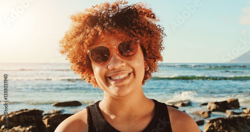  Woman, curly hair and sunglasses , smiling against a backdrop of ocean waves and rocky coastline under a clear sky. High quality photo