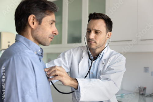 Middle-aged 40s cardiologist using stethoscope to listen to male patient heartbeat, assessing cardiac or respiratory health during medical examination in consulting room. Cardiology, professional care
