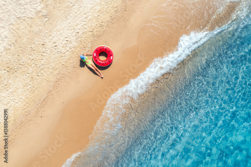 Aerial view of a lying young woman with red swim ring on sandy beach and blue sea at sunset in summer. Tropical landscape with girl, ocean with clear water, waves. Top view. Sardinia island, Italy