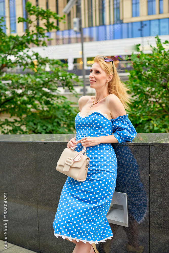 Fototapeta premium A woman in a blue polka dot dress is standing in front of a building. She is holding a white purse and smiling