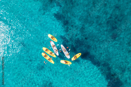 Aerial view of yellow kayaks in blue sea at sunset in summer. People on floating canoes in clear azure water. Sardinia island, Italy. Tropical landscape. Sup boards. Active travel. Top view from drone