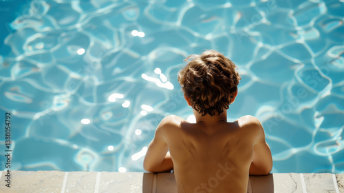 A boy leans against the edge of a sunlit swimming pool, gazing into the reflective blue water.