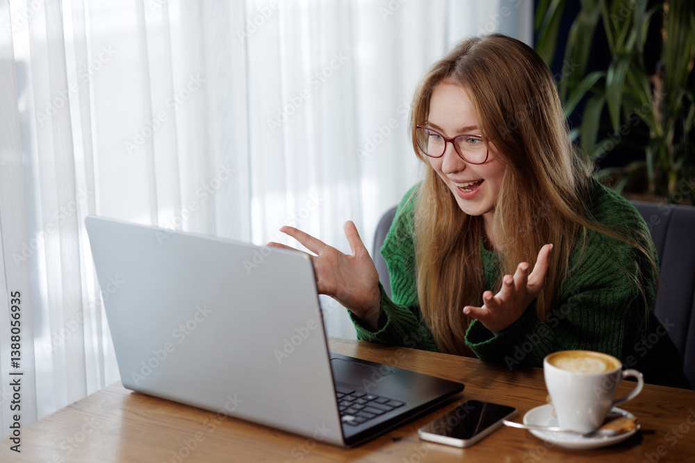 Happy woman with glasses celebrates while using laptop during online conversation in cafe.