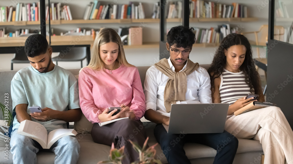 Fototapeta premium Modern study session. Diverse group of multiethnic teen students sit in row on sofa at campus library use mix of gadgets paper materials for learning self training involved in personal education tasks