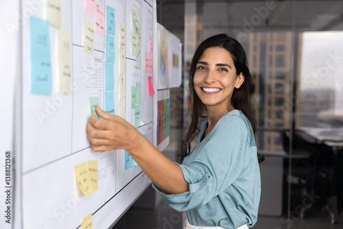 Happy beautiful Hispanic businesswoman working on project management strategy, planning business process alone, sticking paper notes at Kanban board, looking at camera, smiling