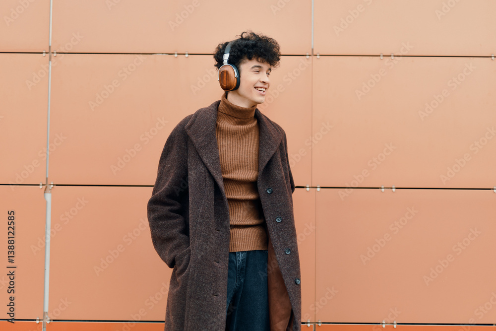 Fashionable young man in headphones and a brown coat smiling in front of an orange wall with hands in pockets