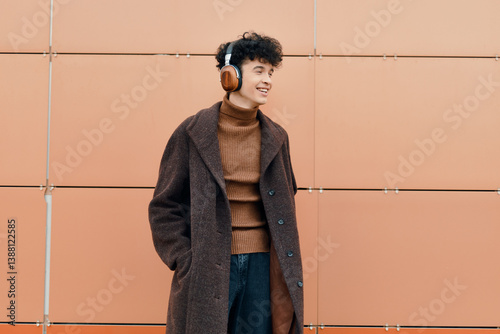 Obraz na płótnie Fashionable young man in headphones and a brown coat smiling in front of an orange wall with hands in pockets w salonie