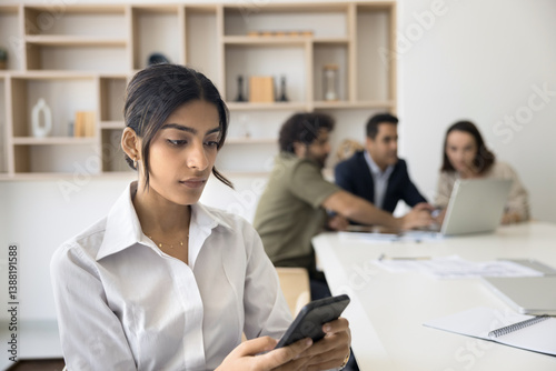 Indian office employee sit at desk using phone, teammates on background engaged in discussion and laptop usage. Modern tech usage for easy, comfortable workflow, ai, innovation, new business app users