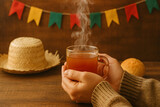 Hands holding a steaming cup of quentão, traditional Brazilian hot drink, with straw hat and colorful Festa Junina bunting