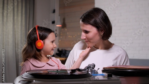 Young girl in headphones smiles, touching her mother's face as they listen to music on a turntable at home. The cozy atmosphere creates a special moment of togetherness