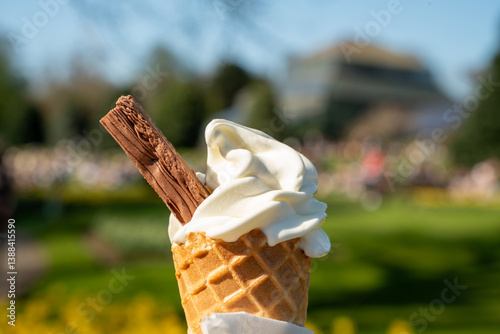 Close-up of a classic vanilla soft-serve ice cream cone with a chocolate flake held in a park on a sunny spring day.