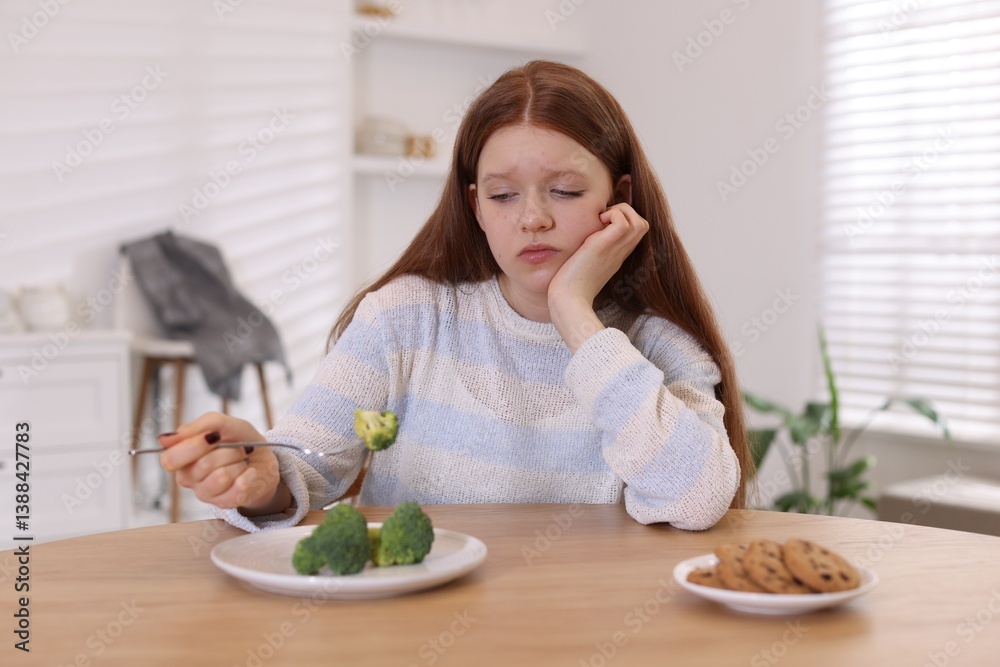 Sad teenage girl with broccoli and cookies at wooden table. Eating disorder Sad teenage girl with broccoli and cookies at wooden table. Eating disorder