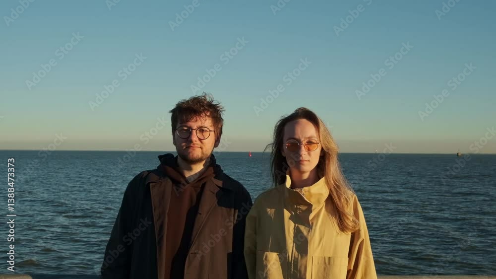 A young couple, a guy and a girl, are standing still, looking directly at the camera. A gentle wind is blowing around them. Behind them are the sea and the horizon.