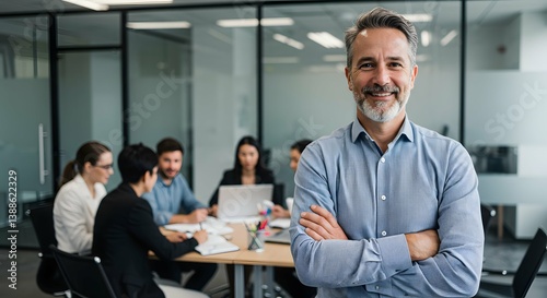 A confident man smiles with arms crossed in a modern office, while a diverse team collaborates around a table in the background during a business meeting.