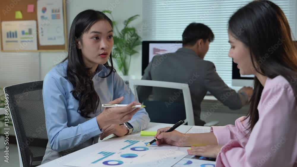Two women are sitting at a table with papers and pens, discussing something. Scene is serious and focused, as the women are engaged in a conversation and working on a project