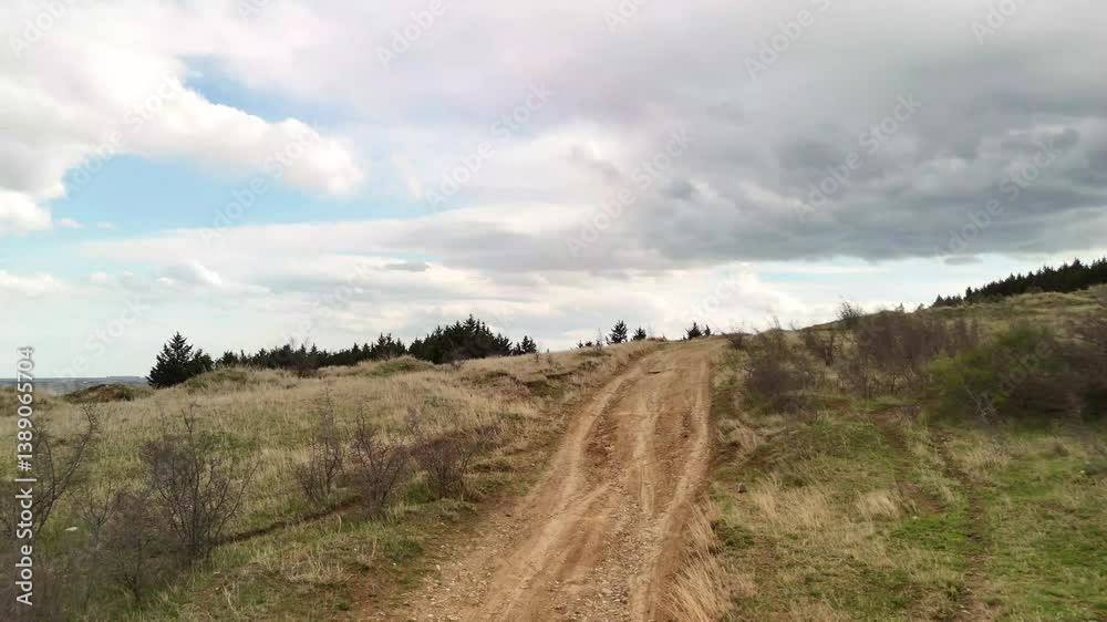 Picturesque countryside landscape featuring a winding dirt road ascending a hill towards a distant forest, under a dynamic sky with a mix of white and dark clouds Picturesque countryside landscape featuring a winding dirt road ascending a hill towards a distant forest, under a dynamic sky with a mix of white and dark clouds