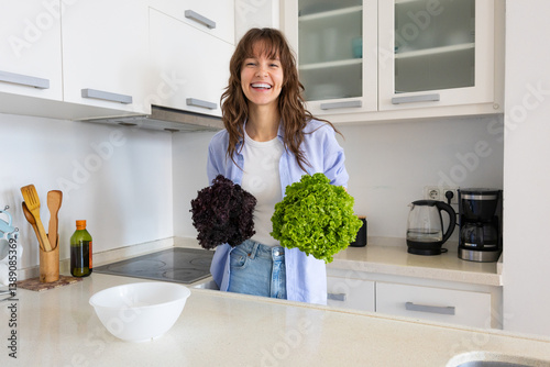 Playful beautiful curly brunette holding two colorful salads, smiling and having fun in the kitchen at home